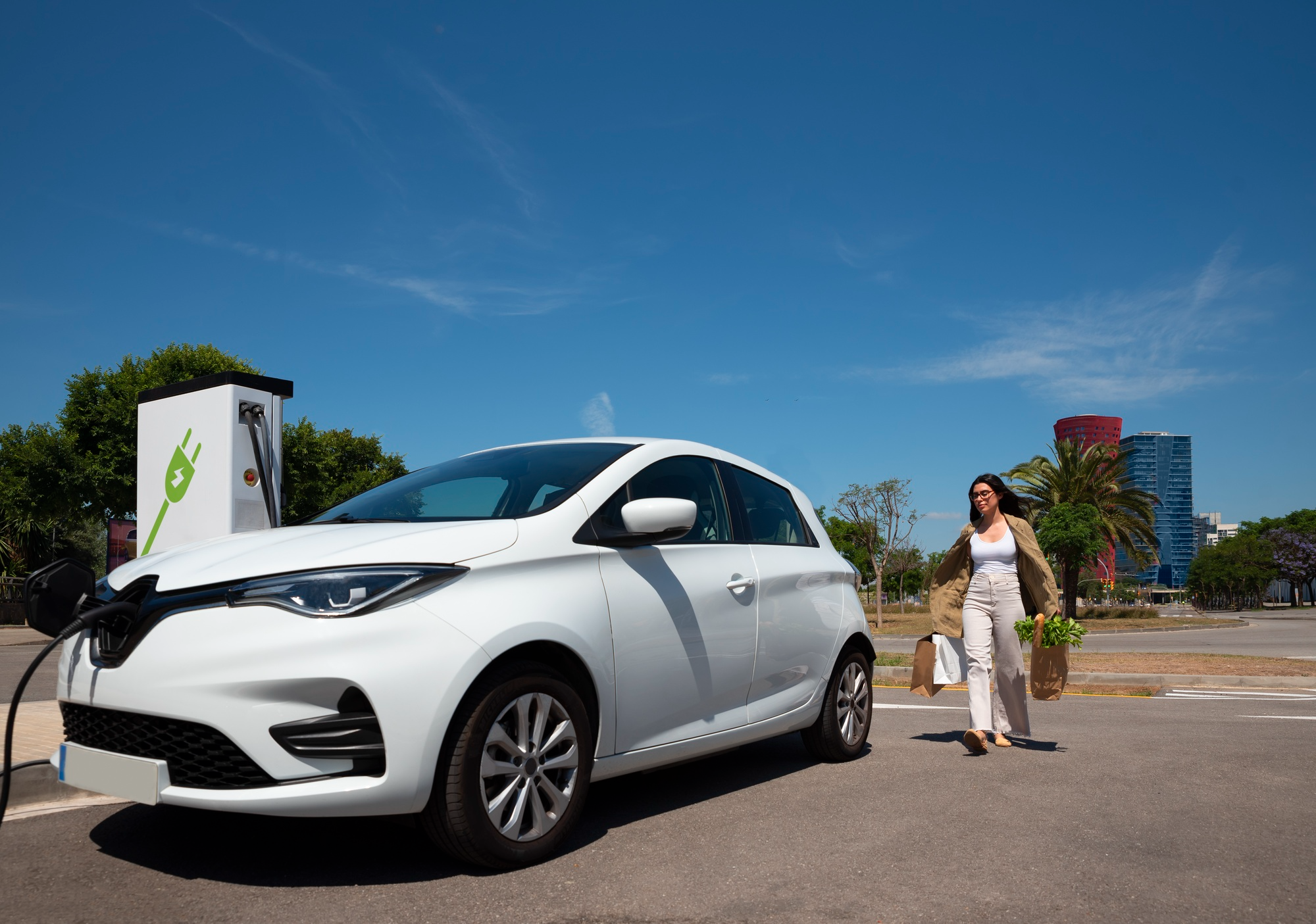 Woman standing next to her electric vehicle at a charging station, holding groceries.